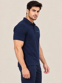 A man modeling a NEAVY BLUE short-sleeve polo shirt with a small white logo on the chest, paired with black jogger pants and white sneakers, against a solid beige studio background.