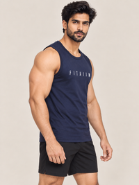 A muscular man wearing a navy blue sleeveless tank top with the word 'FITAIIM' printed in white across the chest, paired with black athletic shorts, posed against a solid light beige background.