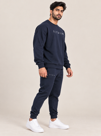 A man modeling a navy blue sweatshirt and jogger pants set with white sneakers, standing against a solid beige studio background.