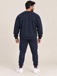 A man viewed from the back wearing a matching navy blue sweatshirt and jogger pants set, paired with white sneakers, against a solid light beige studio background.
