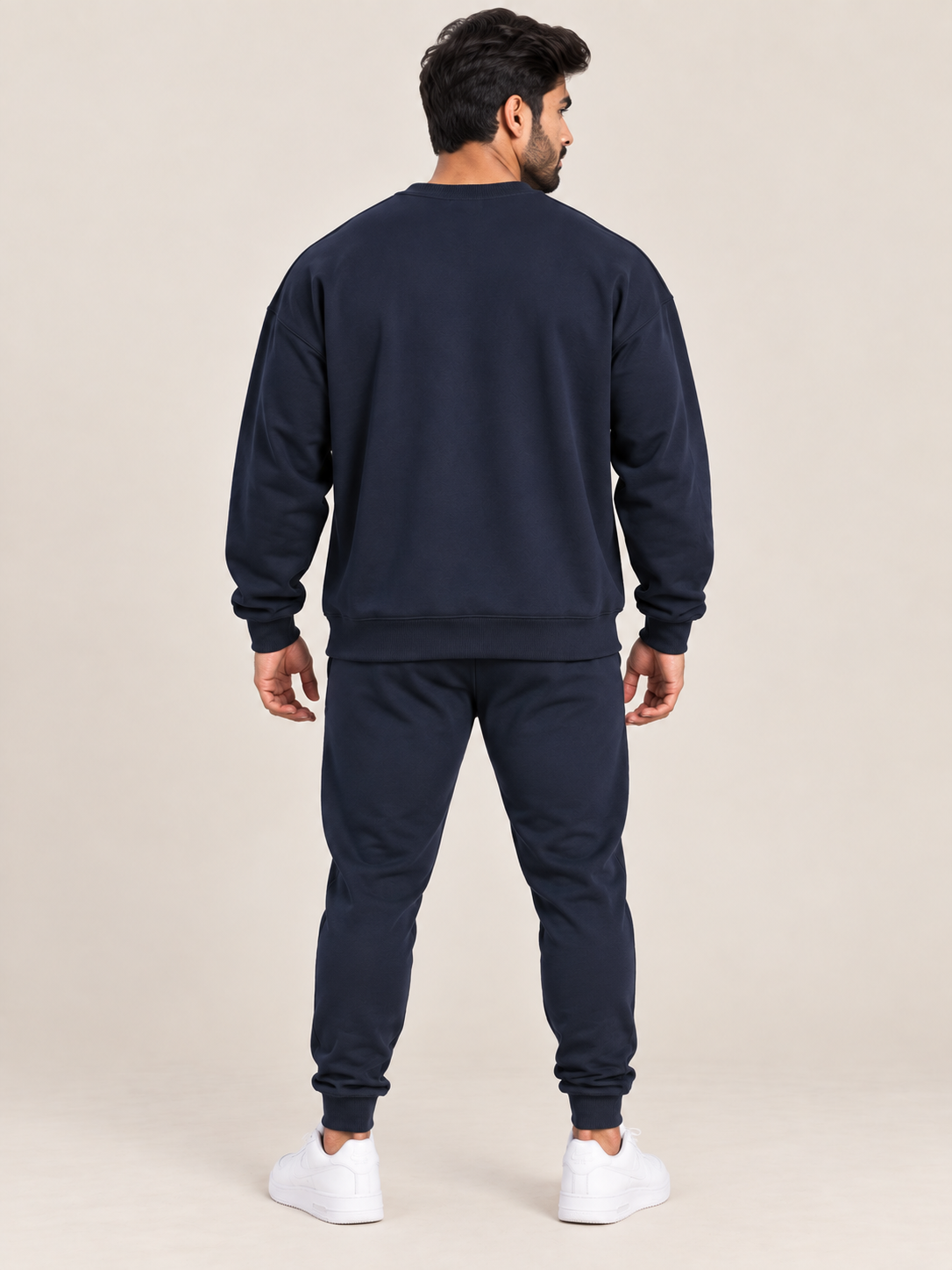 A man viewed from the back wearing a matching navy blue sweatshirt and jogger pants set, paired with white sneakers, against a solid light beige studio background.