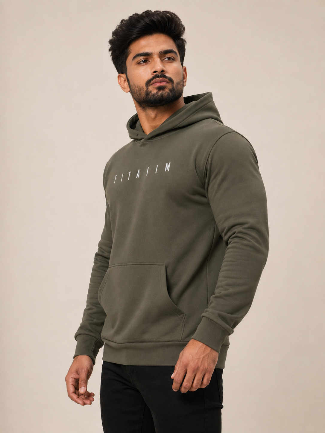 A man wearing an olive green pullover hoodie with 'FITAIM' printed in white across the chest, paired with black pants, set against a plain beige studio background.
