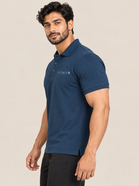 A man wearing a navy blue short-sleeve polo shirt with 'FITAIIM' printed on the chest, paired with black trousers, set against a plain beige studio background.