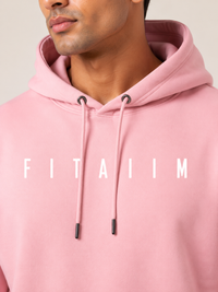 A close-up of a man wearing a light pink hoodie with the word 'FITAIIM' printed in white across the chest. The shot focuses on the upper torso, neck, and lower face of the model.