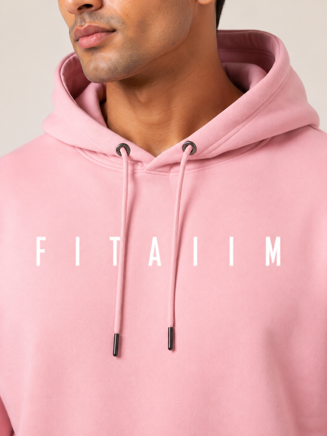 A close-up of a man wearing a light pink hoodie with the word 'FITAIIM' printed in white across the chest. The shot focuses on the upper torso, neck, and lower face of the model.
