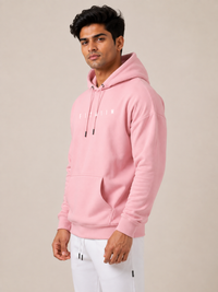 A man wearing a light pink pullover hoodie with white drawstrings and a small white 'STAIN' graphic on the chest, paired with white sweatpants, set against a plain beige studio background.