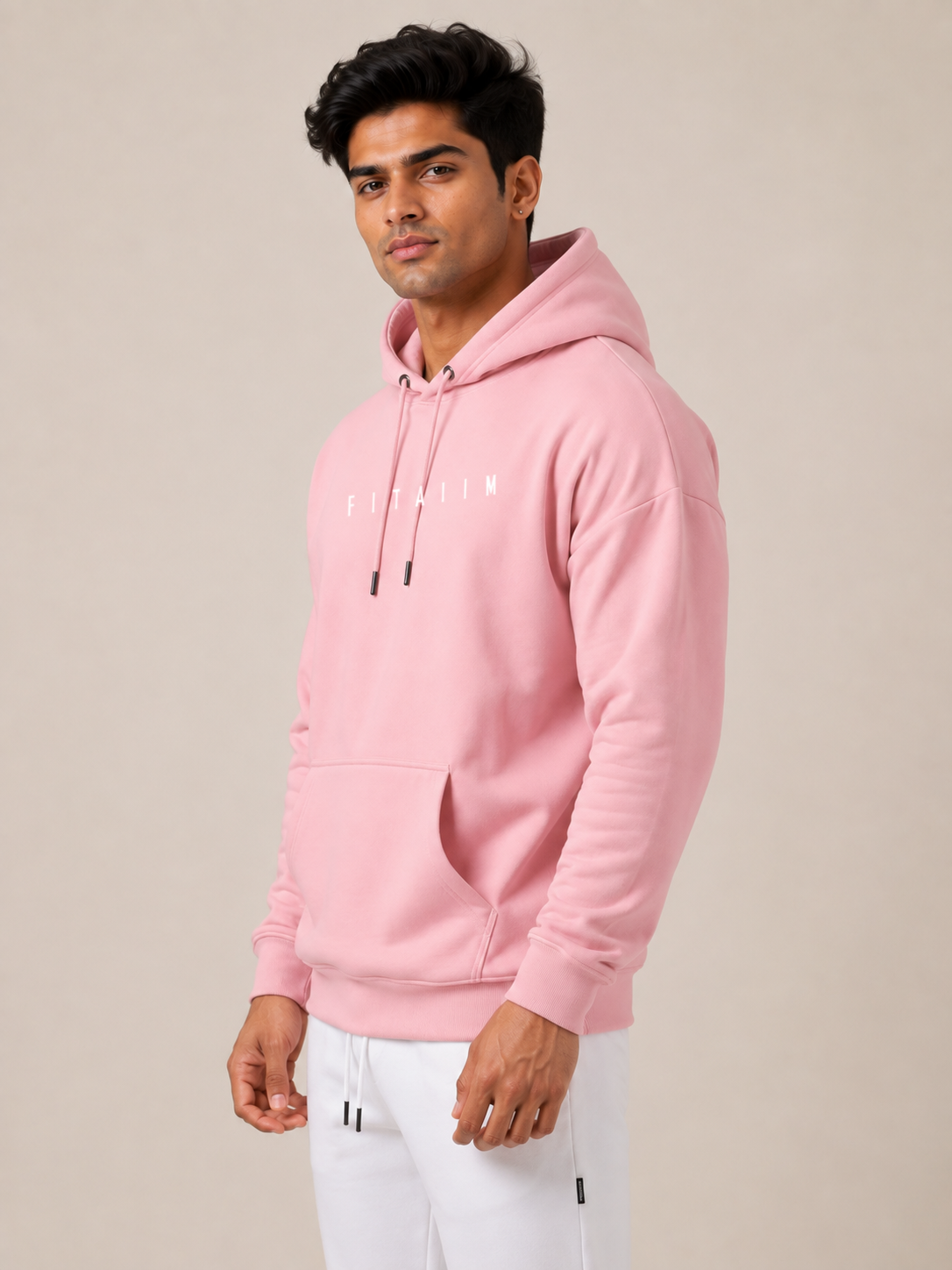 A man wearing a light pink pullover hoodie with white drawstrings and a small white 'STAIN' graphic on the chest, paired with white sweatpants, set against a plain beige studio background.