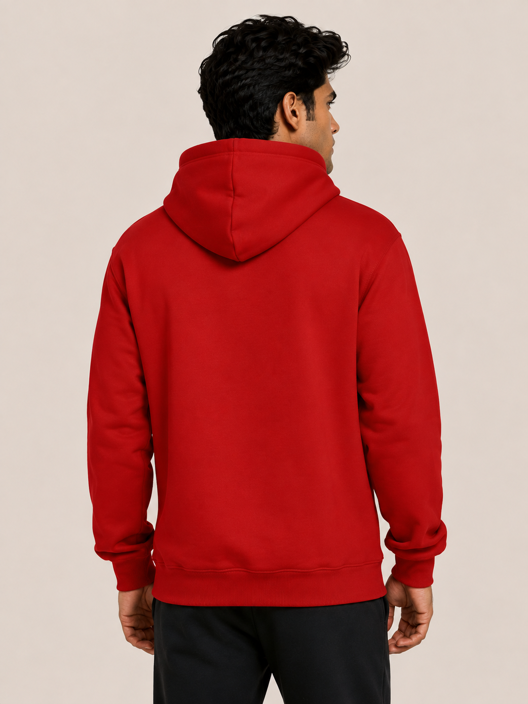 A man viewed from the back wearing a solid red pullover hoodie and black pants, standing against a plain beige studio background.