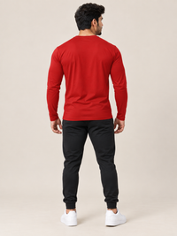 A man shown from the back wearing a bright red long-sleeve crewneck t-shirt, black jogger pants, and white sneakers, standing against a solid light beige background.