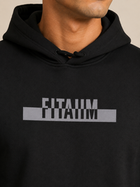A close-up of a person wearing a black pullover hoodie with a gray graphic print on the chest that reads 'FITAIIM' in a bold, blocky font.