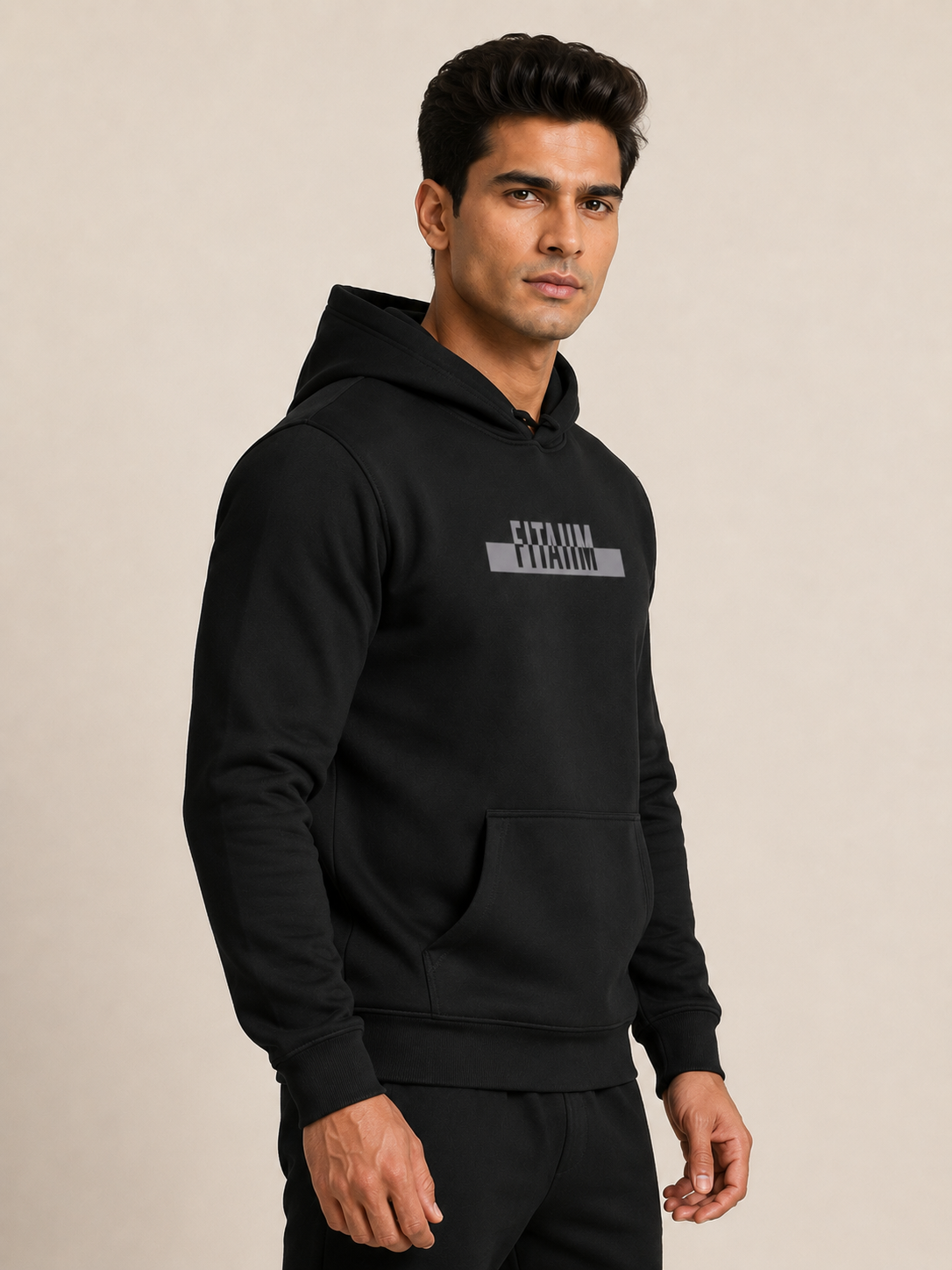 A man wearing a black pullover hoodie with a gray 'FITAIIM' graphic on the chest, paired with black pants, photographed against a neutral beige studio background.