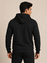 A man viewed from the back wearing a plain black pullover hoodie and matching black pants against a warm beige studio background.