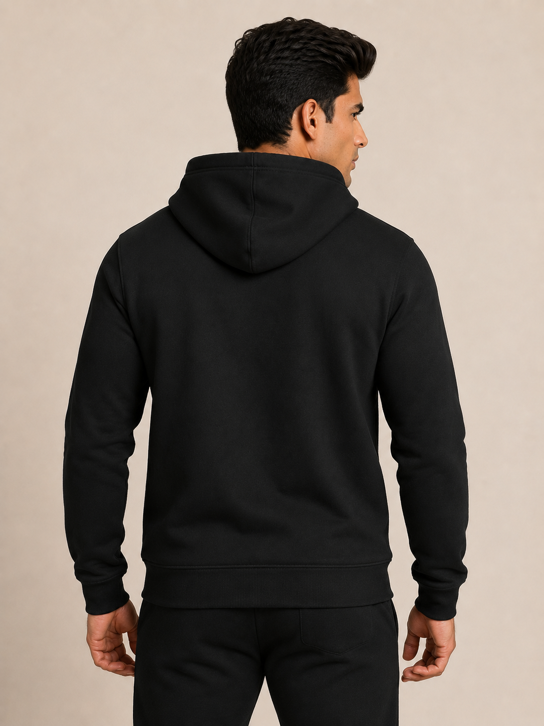 A man viewed from the back wearing a plain black pullover hoodie and matching black pants against a warm beige studio background.