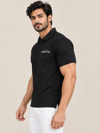 A man wearing a black short-sleeve polo shirt with a small white logo on the left chest, paired with white pants, photographed against a plain beige studio background.