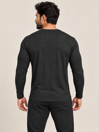 A man shown from the back wearing a dark charcoal gray long-sleeve crewneck shirt and black pants, standing against a solid light beige background.