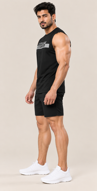 A muscular man modeling a black sleeveless athletic tank top and matching black shorts, paired with white sneakers, against a solid light beige background.