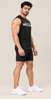 A muscular man modeling a black sleeveless gym tank top with a gray graphic and black athletic shorts, paired with white sneakers, against a solid beige background.