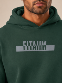A close-up of a man wearing a dark green hoodie with a gray graphic on the chest that reads 'FITAIIM.