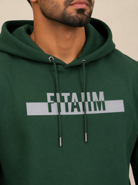 A close-up of a man wearing a dark green hoodie with the name 'FITAIIM' printed in grey capital letters across the chest. The shot focuses on the chest area, showing the drawstrings and the text clearly against a neutral beige background.