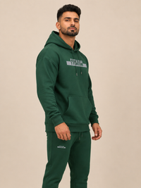 A man wearing a dark green pullover hoodie with 'FITAIIM' printed in grey across the chest, paired with green jogger, against a solid light beige background.