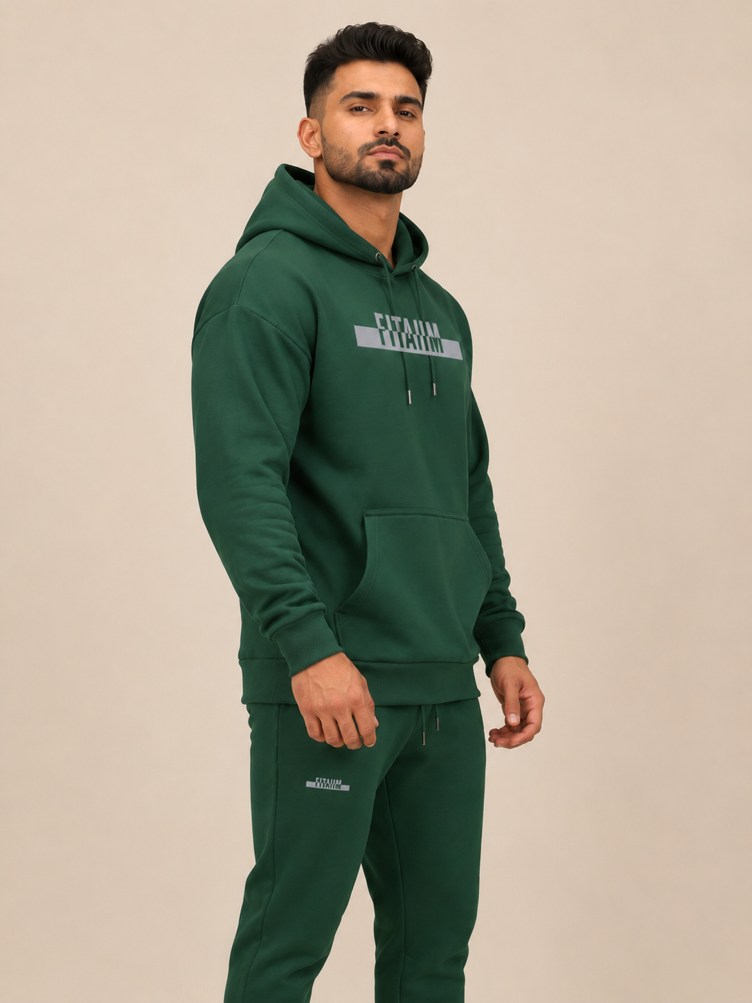 A man wearing a dark green pullover hoodie with 'FITAIIM' printed in grey across the chest, paired with green jogger, against a solid light beige background.