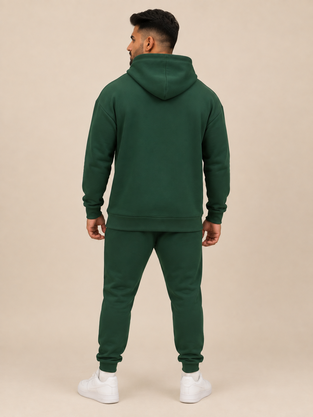 A man shown from the back wearing a dark forest green pullover hoodie and green jogger, standing against a solid beige studio background.