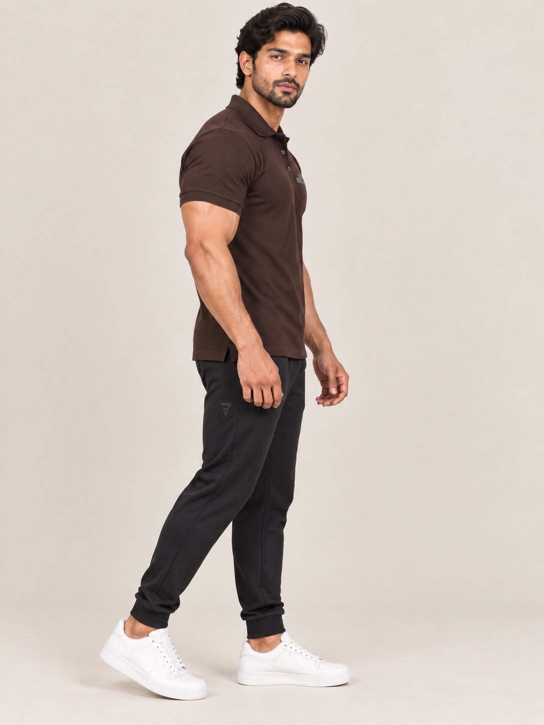A man modeling a dark brown short-sleeve polo shirt, black jogger pants, and white sneakers against a solid light beige background.