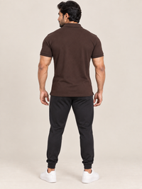A man shown from the back wearing a dark brown short-sleeve polo shirt, black jogger pants, and white sneakers, standing against a plain beige studio background.