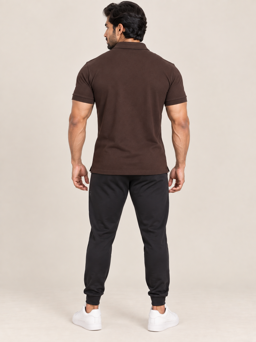 A man shown from the back wearing a dark brown short-sleeve polo shirt, black jogger pants, and white sneakers, standing against a plain beige studio background.
