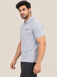 A man wearing a short-sleeve GREY polo shirt with a small gray and white logo on the left chest, paired with black pants, photographed against a plain light beige studio background.
