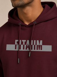 A close-up of a man wearing a maroon hoodie with the name 'FITAIIM' printed in grey capital letters across the chest. The shot focuses on the chest area, showing the drawstrings and the text clearly against a neutral beige background.