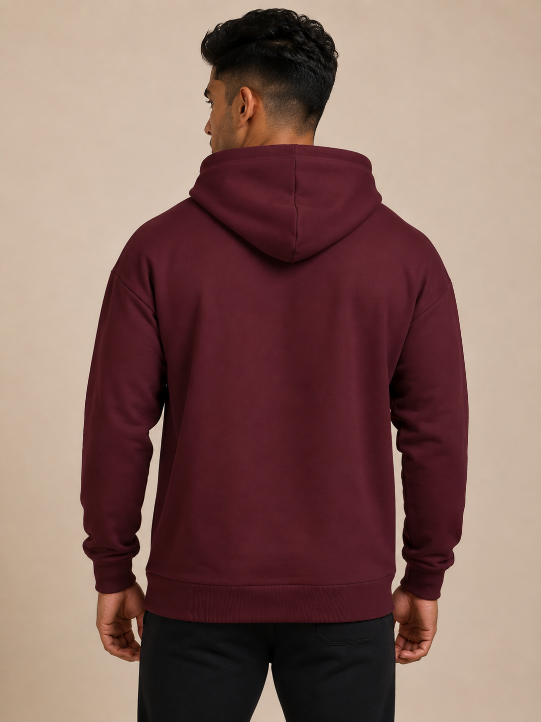 A man shown from the back wearing a maroon pullover hoodie and black jogger, standing against a solid beige studio background.