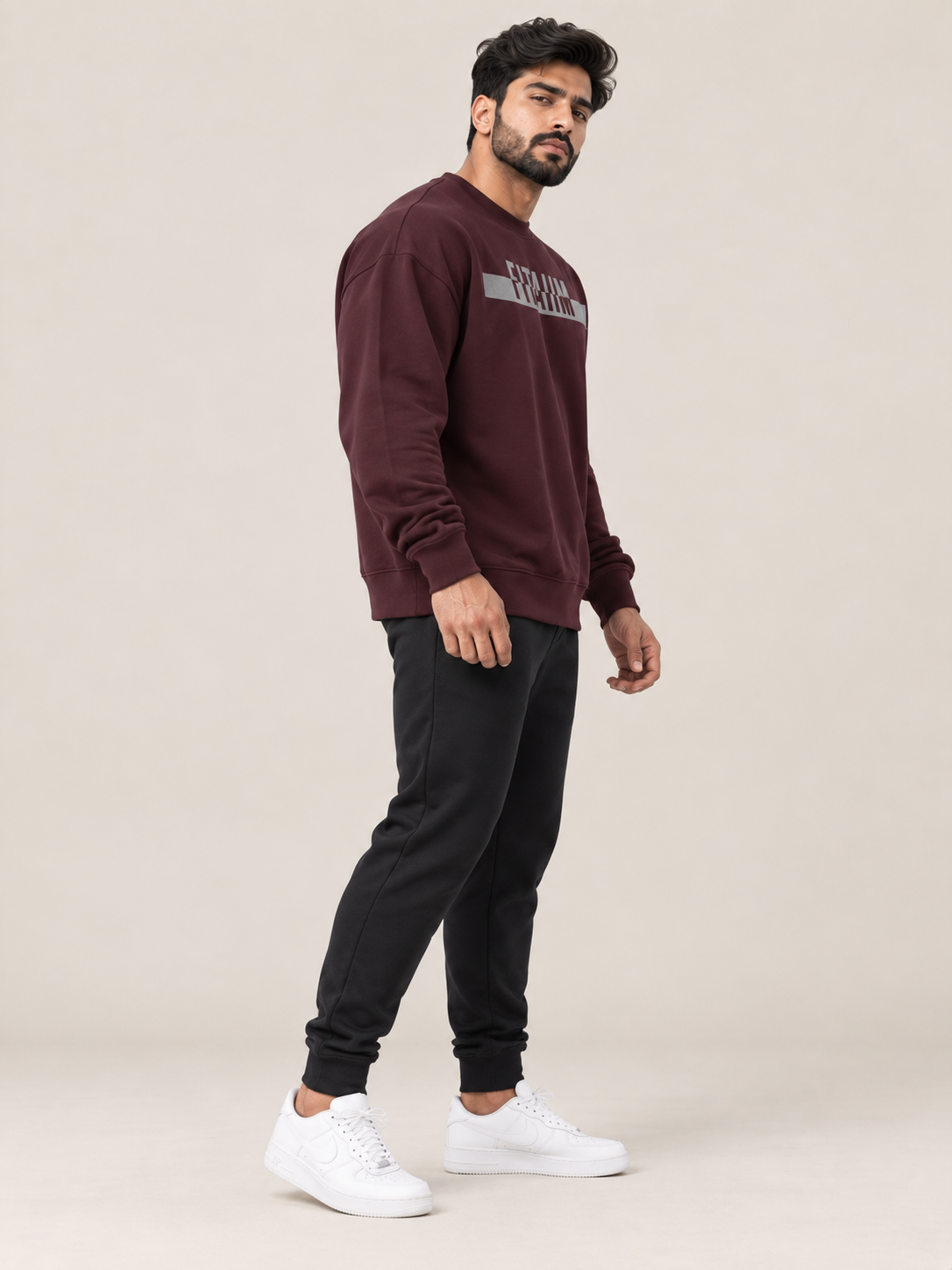 A man modeling a matching maroon sweatshirt and jogger pants set with white sneakers, standing against a solid beige studio background.
