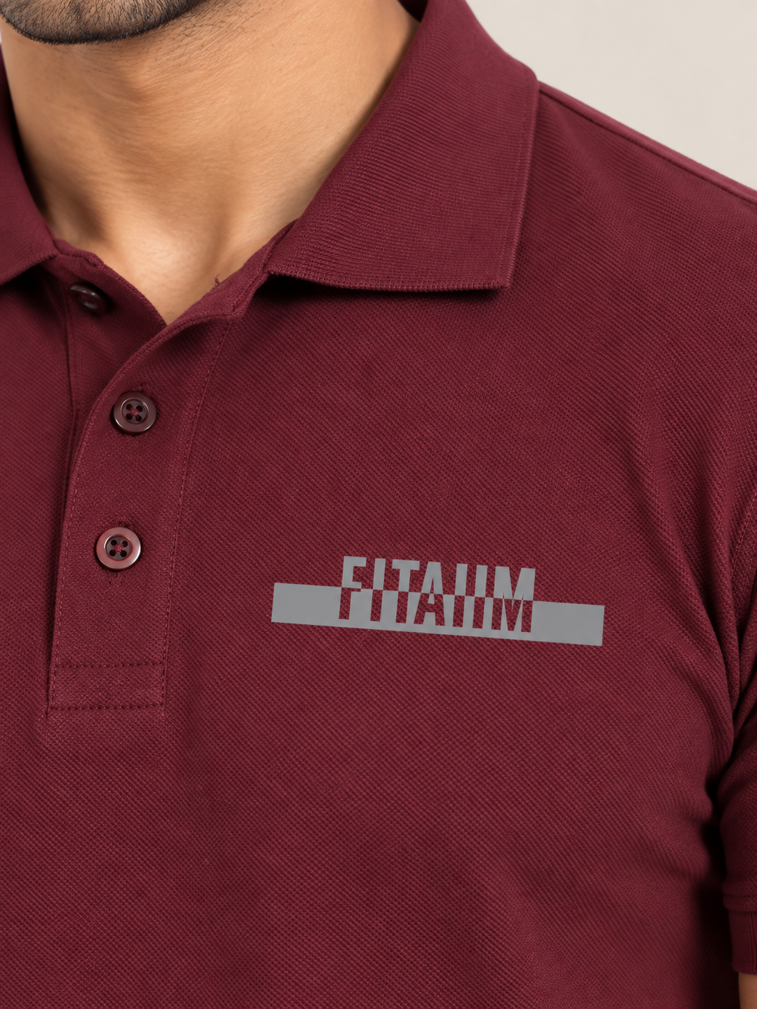 A close-up of a man wearing a MAROON short-sleeve polo shirt with a gray and white 'FITAIIM' graphic printed on the left chest.