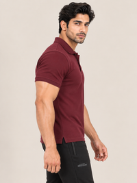 A man wearing a short-sleeve polo shirt in maroon and black pants, standing in profile against a plain beige studio background.