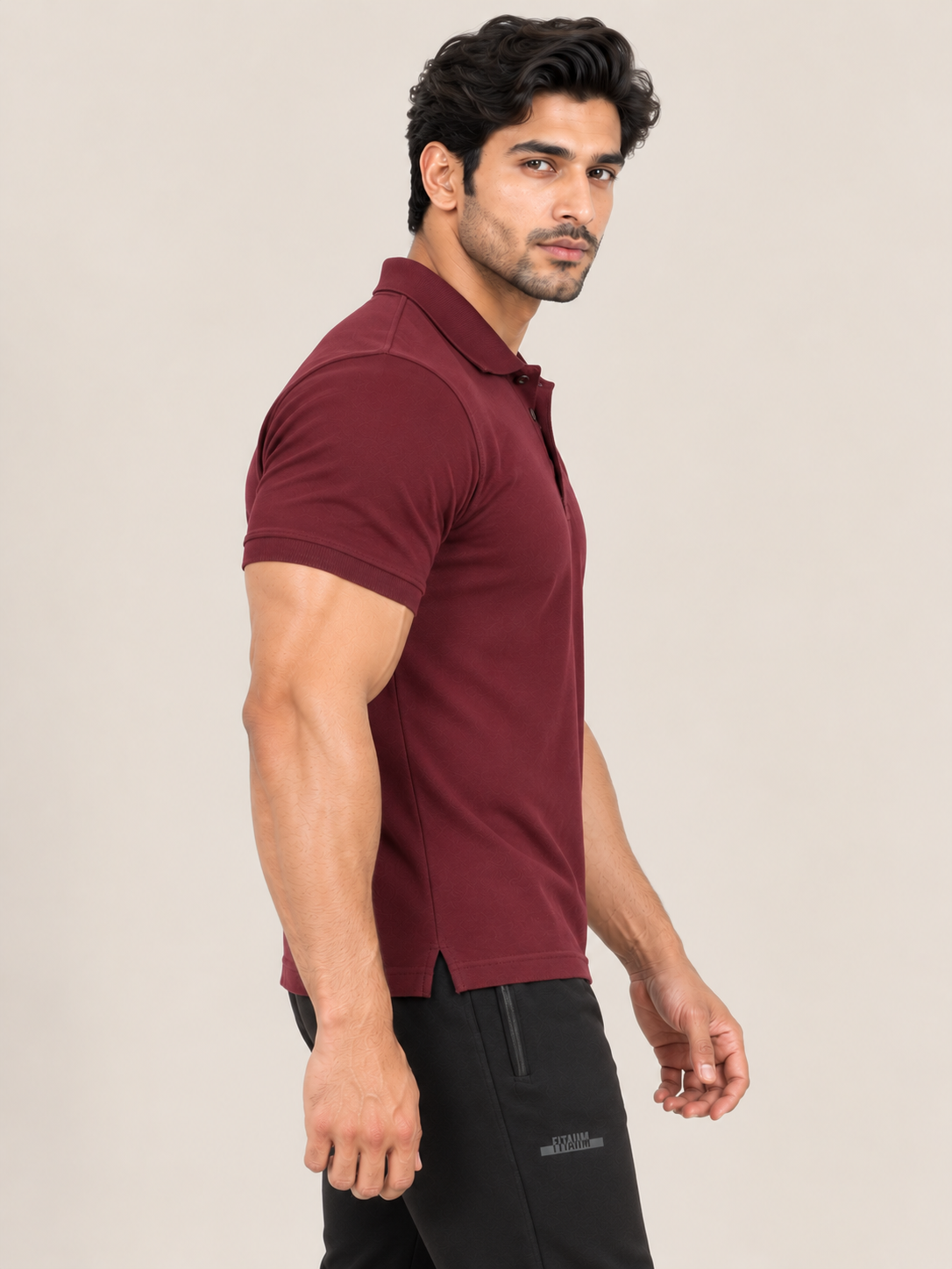 A man wearing a short-sleeve polo shirt in maroon and black pants, standing in profile against a plain beige studio background.