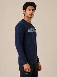A man wearing a navy blue long-sleeve crewneck sweatshirt with a white 'FITAIM' logo across the chest, paired with black sweatpants, set against a plain beige studio background.
