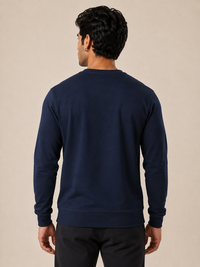 A man viewed from the back wearing a navy blue long-sleeve crewneck sweatshirt and black pants, standing against a solid beige studio background.