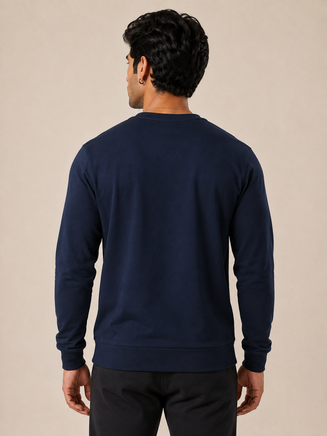 A man viewed from the back wearing a navy blue long-sleeve crewneck sweatshirt and black pants, standing against a solid beige studio background.