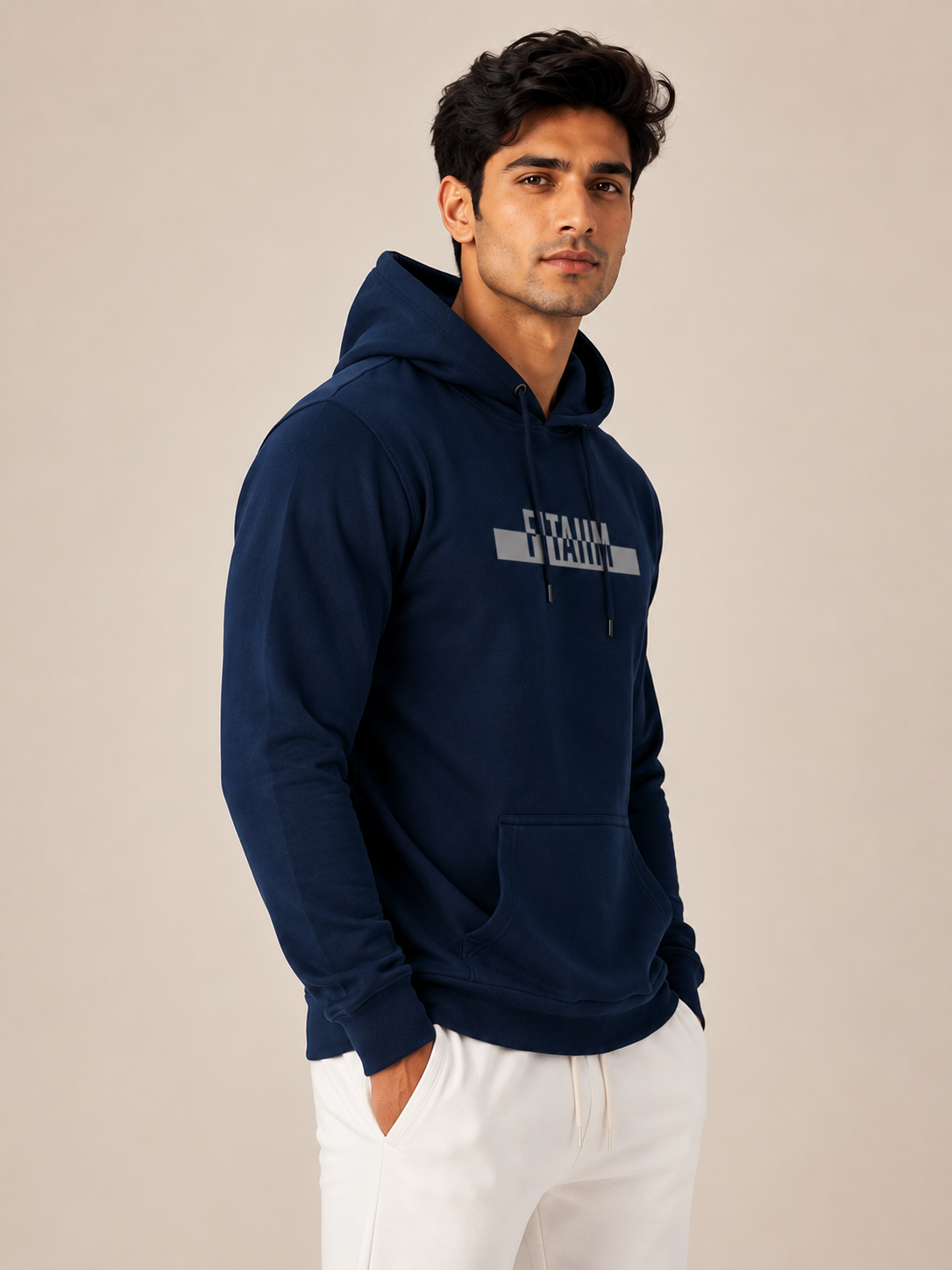 A man wearing a navy blue hoodie with a gray and white graphic on the chest, paired with white pants, standing against a plain beige background.