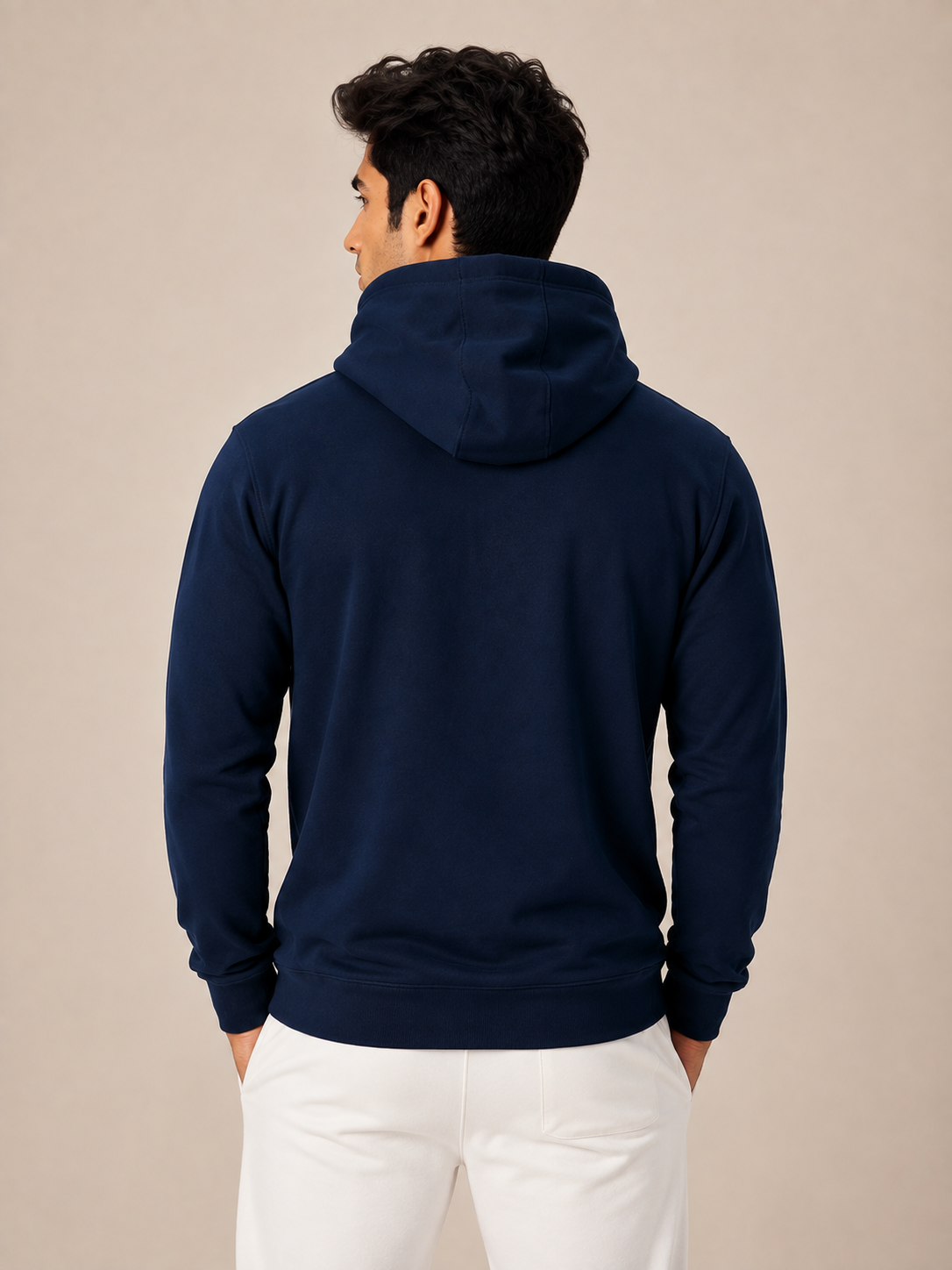 A man viewed from the back wearing a navy blue pullover hoodie and white pants, standing against a plain beige studio background.