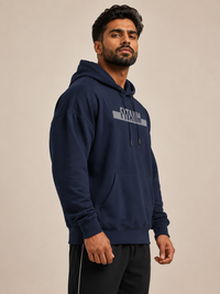A man wearing a navy blue pullover hoodie with a gray graphic text print on the chest and matching black athletic shorts, standing against a plain beige studio background.