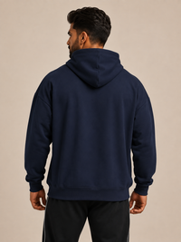 A man viewed from the back wearing a navy blue pullover hoodie and black track pants with white side stripes, set against a plain beige studio background.