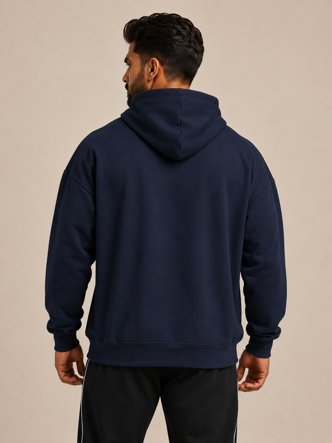 A man viewed from the back wearing a navy blue pullover hoodie and black track pants with white side stripes, set against a plain beige studio background.