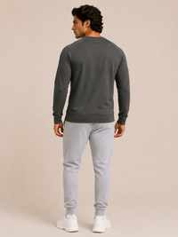 A man shown from the back wearing a dark olive green long-sleeve crewneck sweatshirt, light gray jogger sweatpants, and white sneakers, standing against a solid beige studio background.