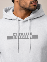 A close-up of a man wearing a light gray heather hoodie with a dark gray graphic print on the chest that reads 'FITAIIM'.