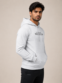 A man wearing a light gray heathered pullover hoodie with a dark gray 'FITAIIM' logo on the chest, paired with black pants, photographed against a solid beige studio background.