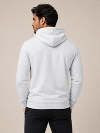 A man viewed from behind wearing a light gray heather pullover hoodie and black pants, standing against a solid beige studio background.