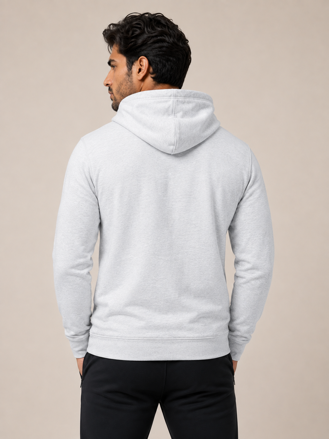 A man viewed from behind wearing a light gray heather pullover hoodie and black pants, standing against a solid beige studio background.