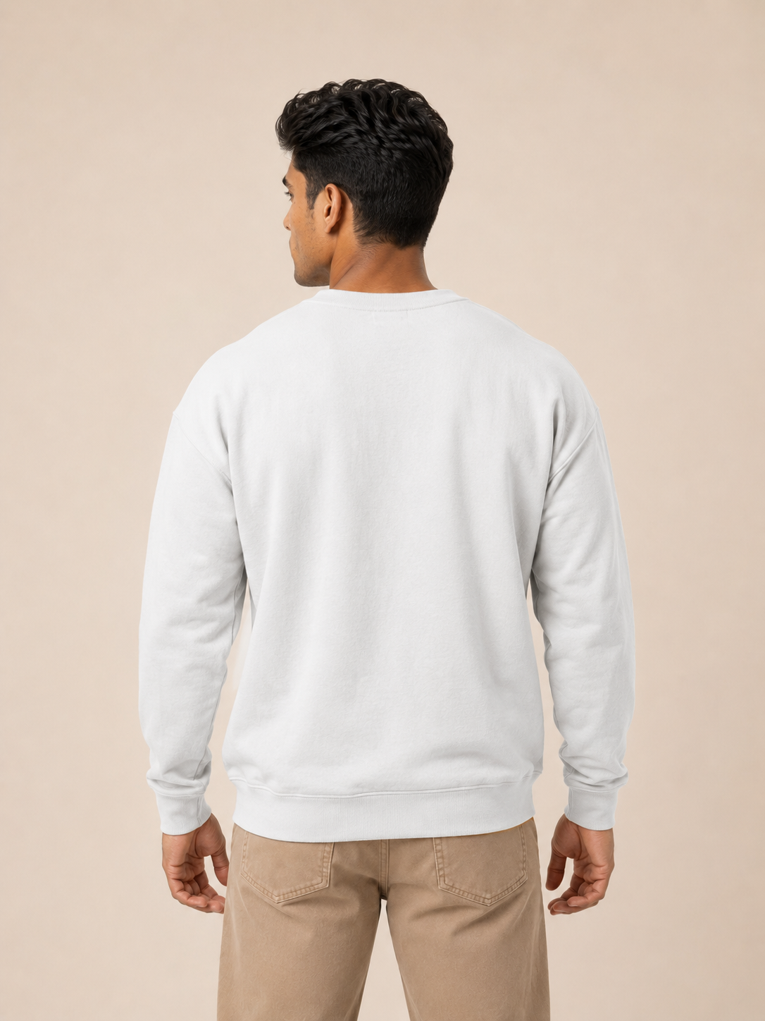 A man viewed from the back wearing a plain white sweatshirt and matching beige pants against a warm beige studio background.